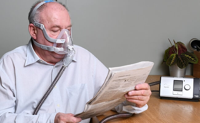 COPD_Patient_mit_Lumis_Therapiegerät_und Full Face Maske liest Zeitung