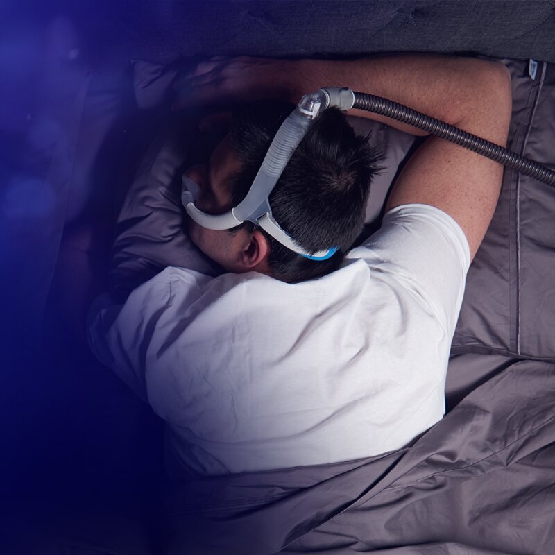 Auf dem Bauch liegender Mann mit einer AirFit N30i CPAP-Maske von Resmed, der neben seiner Partnerin schläft