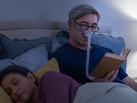 Ein Mann mit Brille liest im Bett, während er eine ResMed AirFit N30 Nasenmaske trägt.