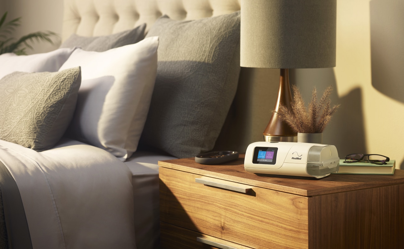 Ein adaptives Servo-Ventilationsgerät AirCurve 11 ASV PaceWave auf einem Nachttisch, daneben eine Lampe, eine Lesebrille und ein Buch.